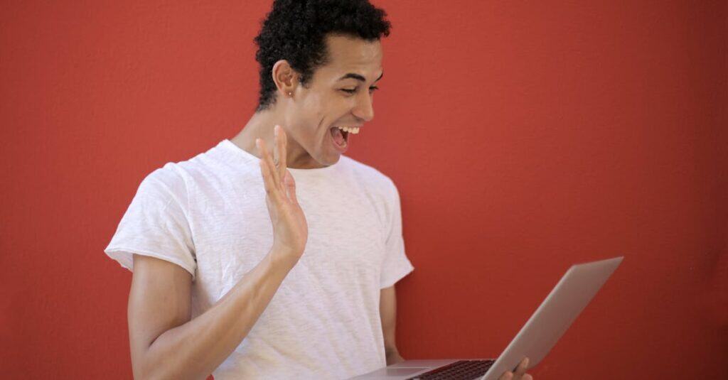 Young man making a cheerful video call on a laptop with a red background, expressing joy and surprise.