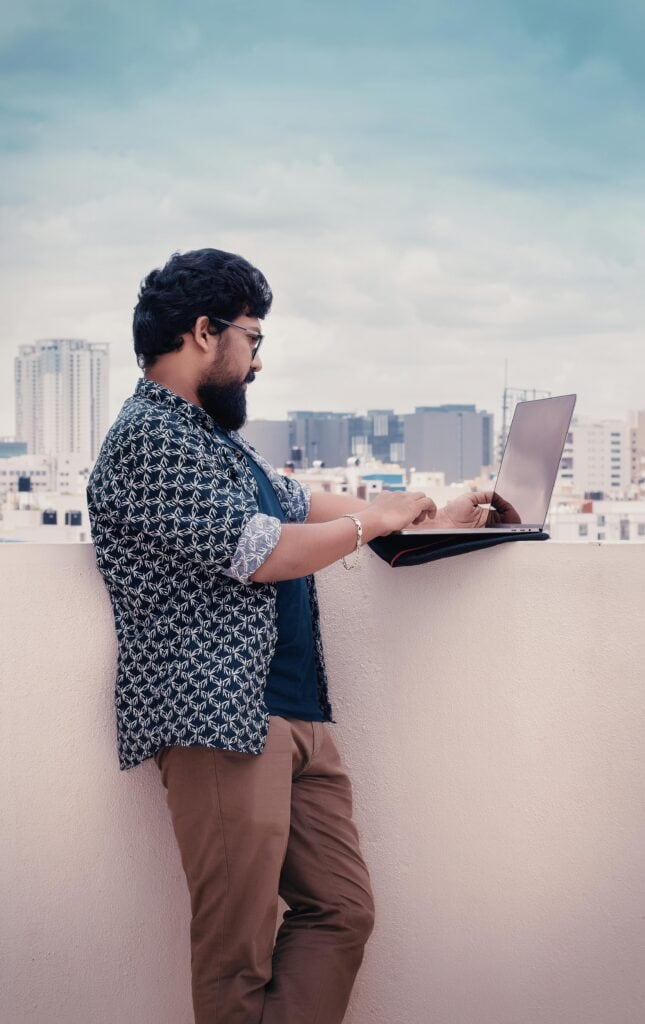 Man working on laptop on a rooftop with Bengaluru city skyline in the background.